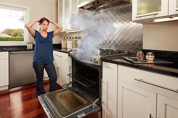 A person in a kitchen beside an open oven with smoke rising above it.